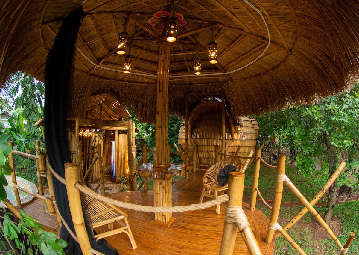 A spacious deck crafted from bamboo showcases natural textures, surrounded by lush greenery. Unique bamboo furniture, including a cozy chair and a circular seating area, invites relaxation. The ceiling is adorned with lanterns, providing soft lighting that enhances the tranquil atmosphere.