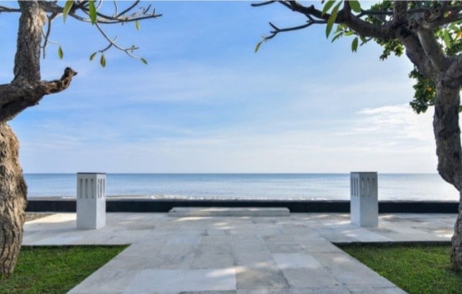 A calm beach view is visible, featuring a sandy shoreline leading to a tranquil sea. Two white stone pillars flank a wide, paved area, providing a transition from the garden to the ocean. Nearby, lush greenery enhances the serene atmosphere.
