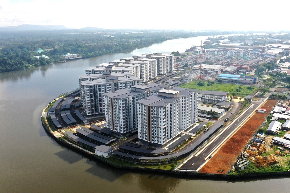 An aerial view captures the riverfront apartment complex, showcasing modern architectural designs. The surrounding landscape includes the river alongside a newly established roadway, with large parking areas visible in front of the buildings, contributing to a sense of accessibility and spaciousness.