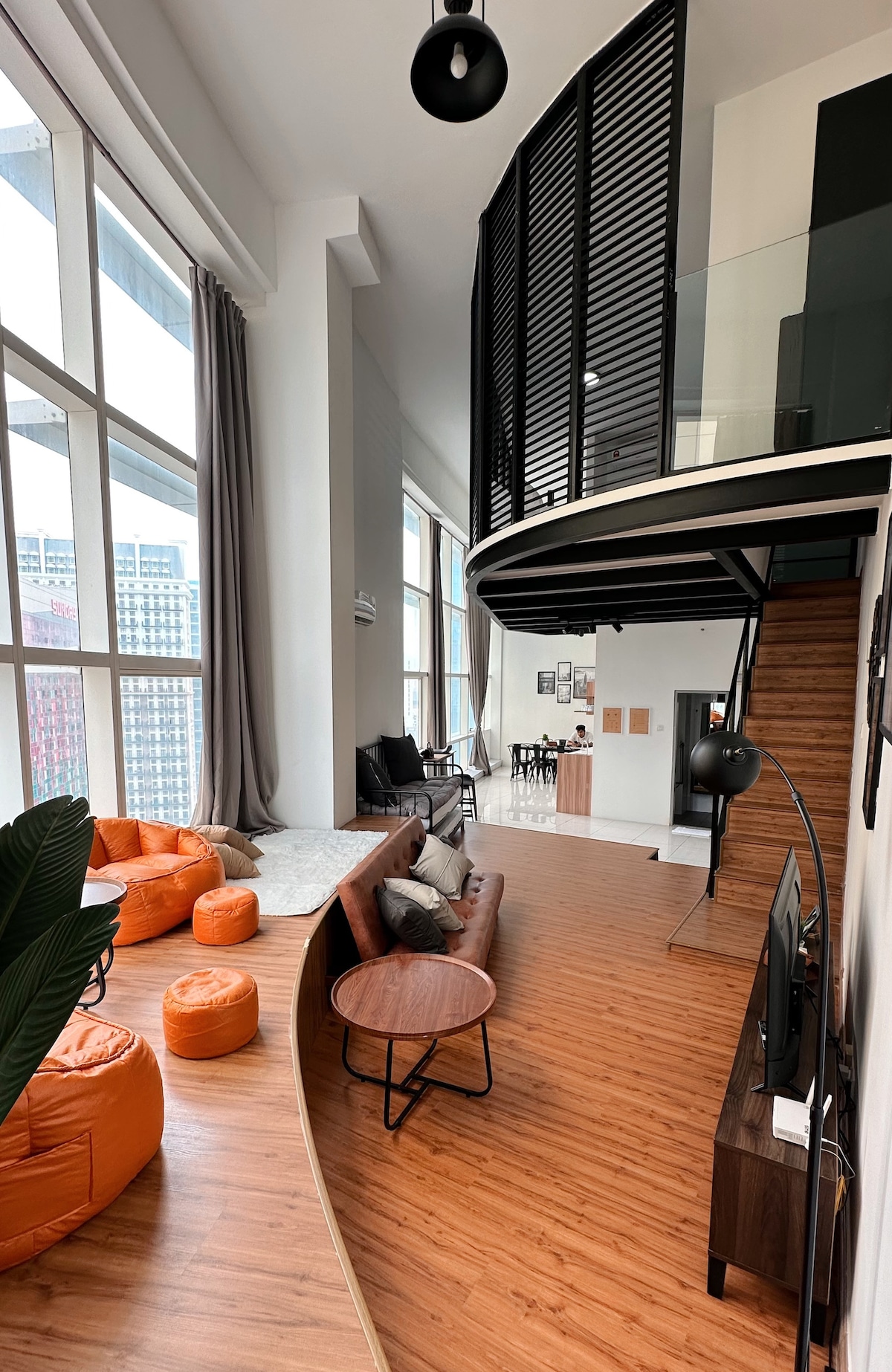 An open-plan living area displays a blend of sleek furnishings and soft textures. A curved wooden seating area is accompanied by orange bean bags, providing a casual vibe. Large windows allow ample natural light, illuminating the space and connecting indoors with the city view.