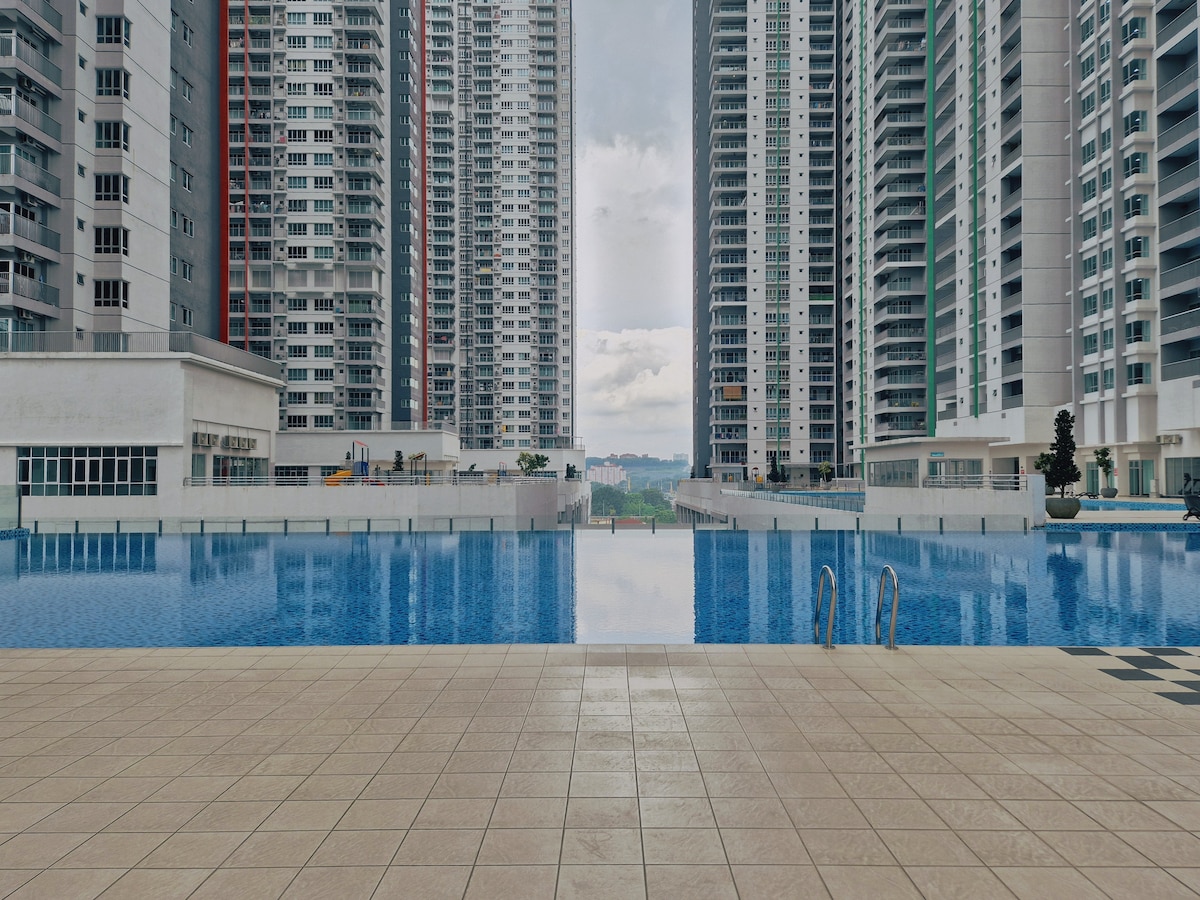 A large swimming pool reflects the surrounding high-rise buildings, creating a striking visual symmetry. The pool area is paved with light-colored tiles, with several steps leading into the water. A tranquil atmosphere is framed by green landscaping in the background.