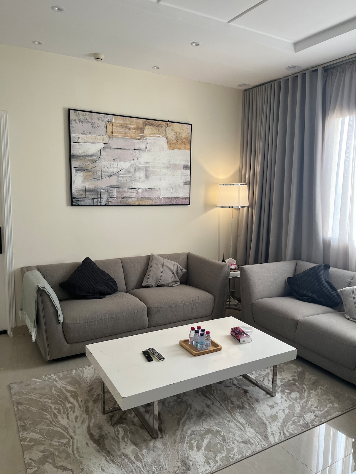 A modern living room is presented with two gray sofas, arranged around a rectangular coffee table. A large piece of abstract wall art is featured above the sofas. Natural light filters through sheer curtains, creating a calm and inviting space.