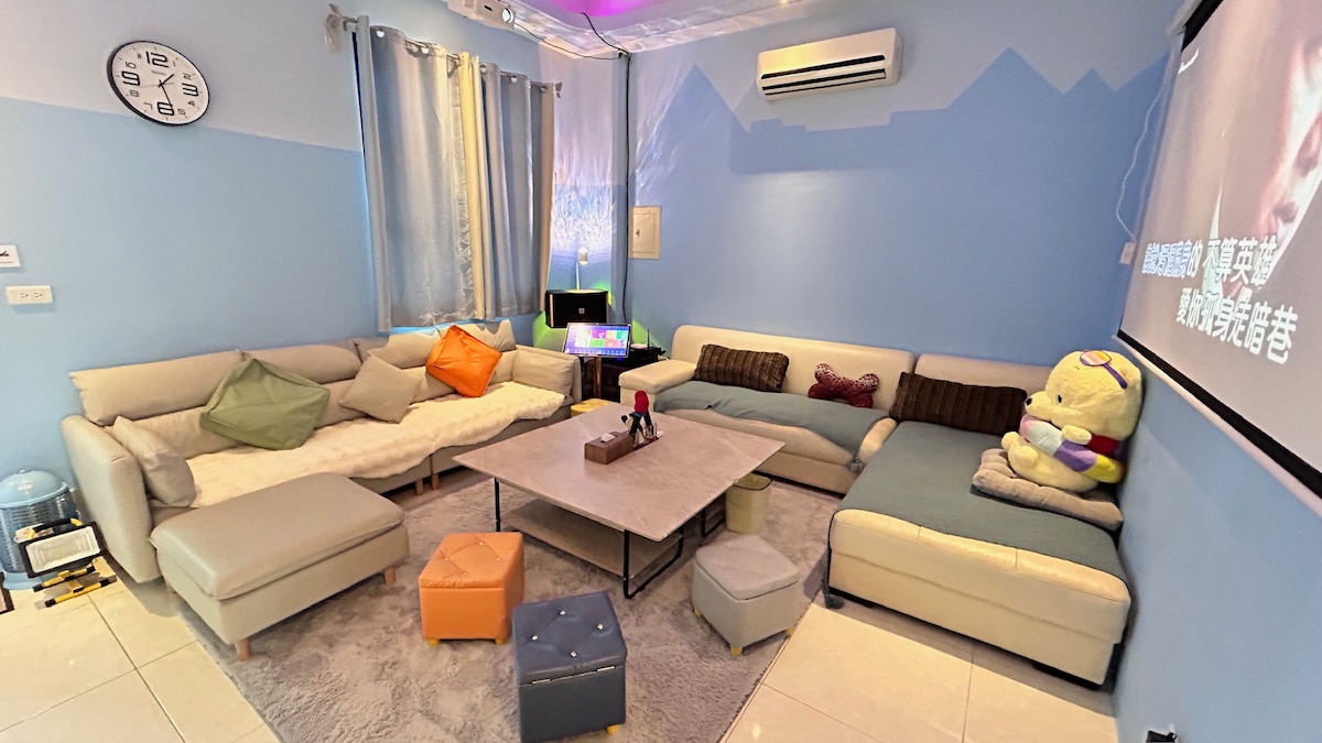 A spacious living area is furnished with a large sectional sofa in neutral tones, accented by colorful cushions. A coffee table is located centrally, while a plush character toy adds a playful touch. Soft lighting enhances the welcoming environment, and a projector screen is mounted on the wall.