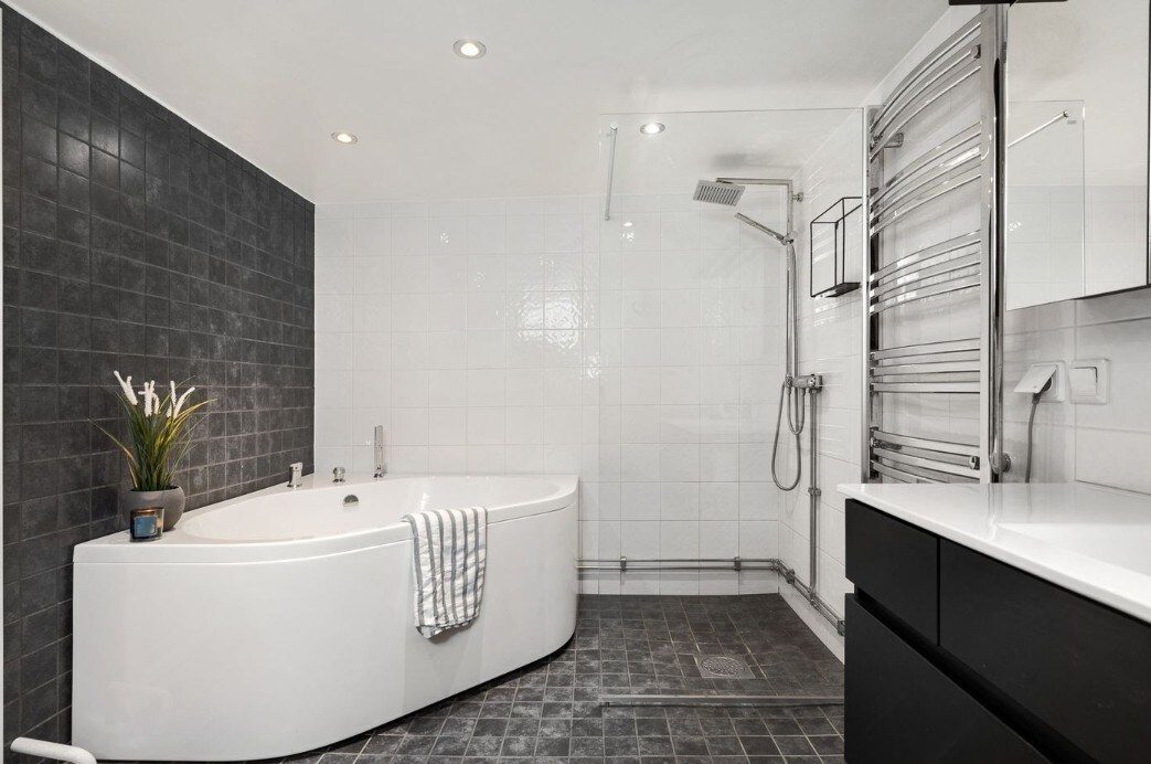 A spacious modern bathroom features a curved white bathtub and a sleek black cabinet. The room is illuminated by recessed lighting, highlighting the contrast between the dark tiles and the white walls. A shower area is equipped with a polished chrome fixture.