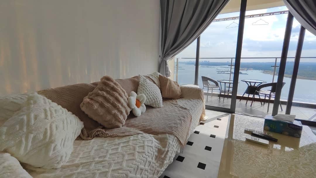 A comfort-focused living area features a plush sofa adorned with decorative pillows and a soft throw. Large glass doors open to a balcony, allowing views of the water and an outdoor seating arrangement with two chairs and a small table.