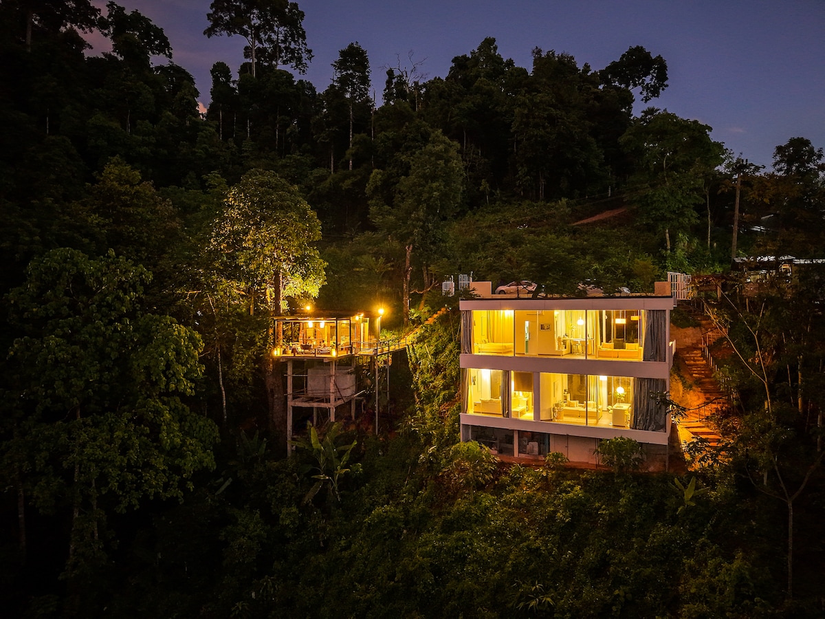 The image displays a modern white glass house nestled in a lush green landscape, illuminated softly by outdoor lights. A separate wooden deck can be seen elevated among the trees, providing a serene atmosphere at twilight.