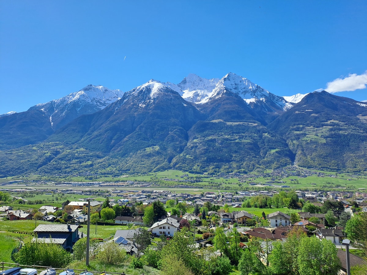 Distant mountains with snow-capped peaks are visible against a clear blue sky. The lush valley below is dotted with small houses and greenery, showcasing a serene landscape that invites relaxation.