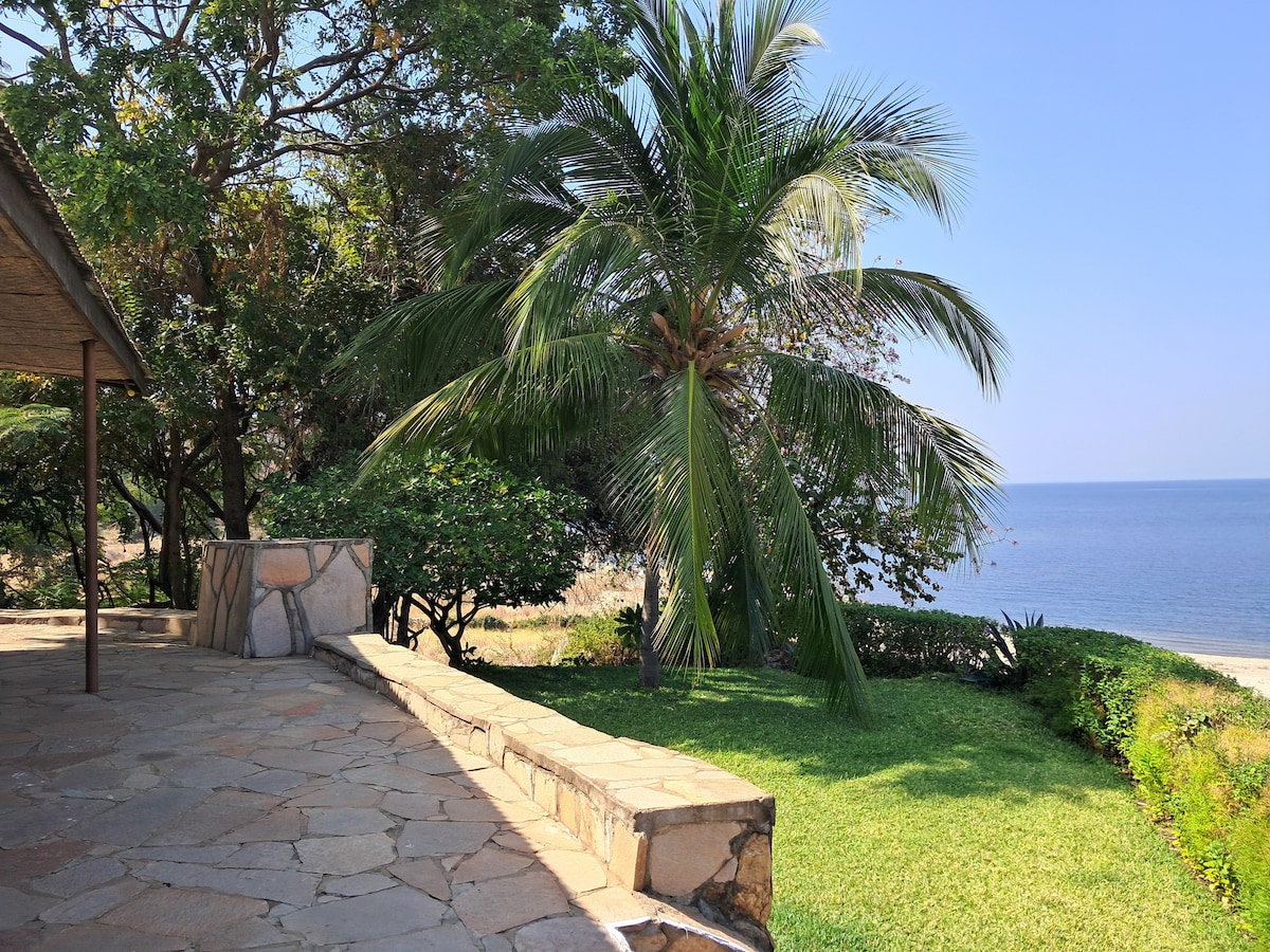 A landscaped outdoor area features a stone wall overlooking the beach and Kariba Lake. Lush greenery and palm trees provide shade, while the calm water sparkles in the sunlight. The scene offers a serene setting for relaxation and enjoyment of nature.