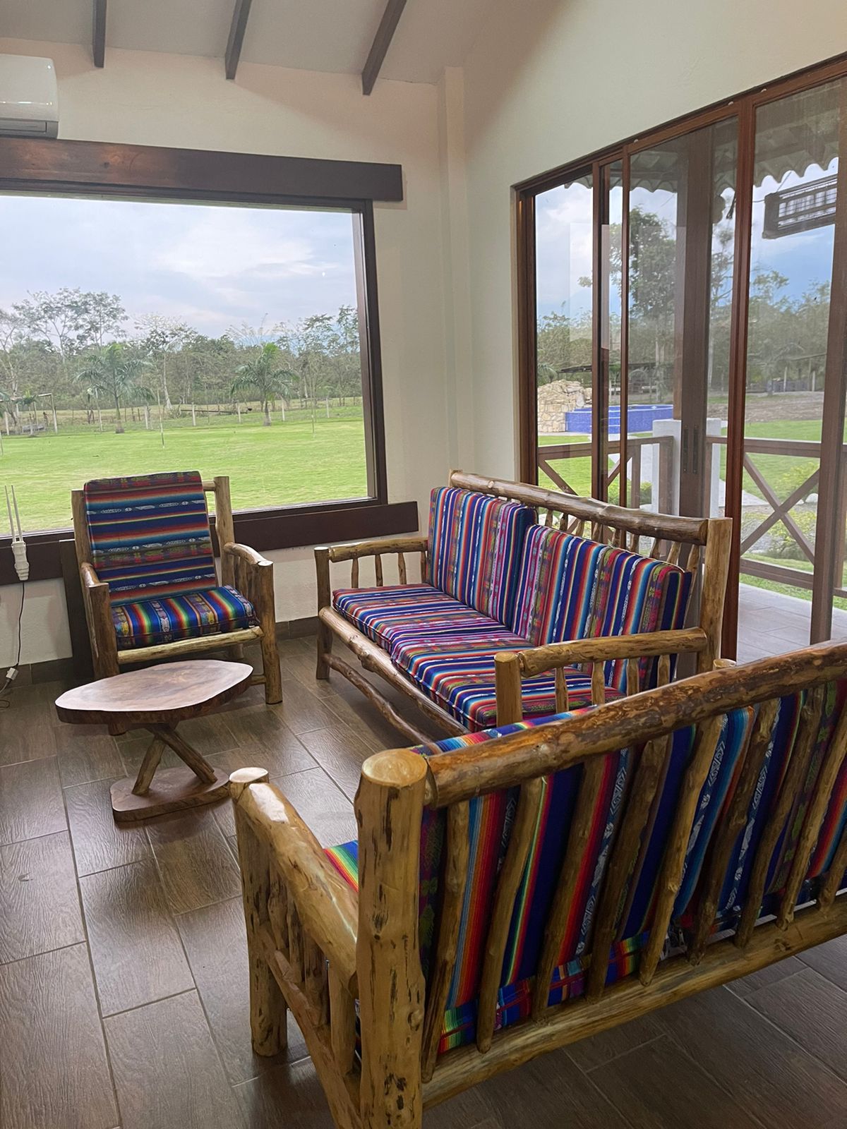 A light-filled seating area features a cozy sofa and armchair with colorful striped upholstery. A wooden coffee table sits in the center, and large windows provide views of the surrounding greenery.