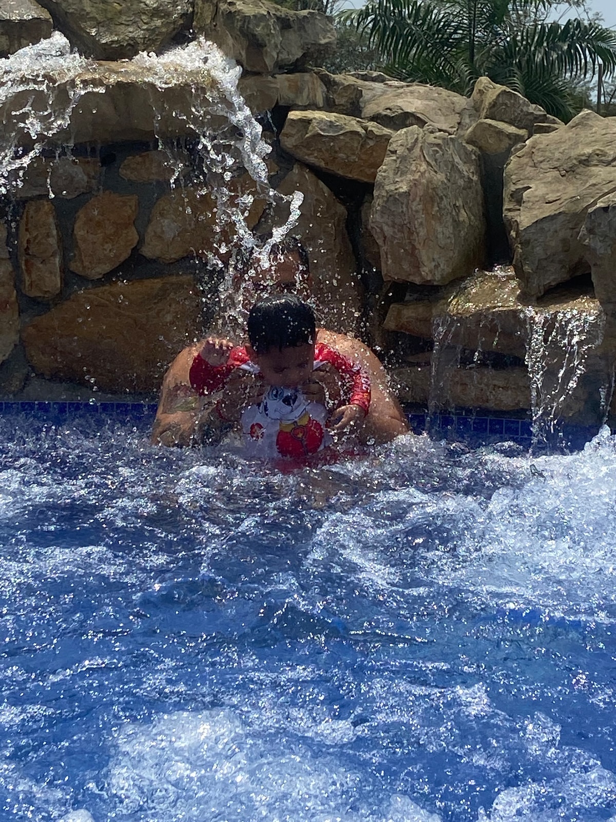 A water play area is depicted, featuring a rocky backdrop with water cascading over stones. In the foreground, splashes of water are visible, indicating fun activities in the pool. Bright sunlight reflects off the water's surface, creating a lively atmosphere.