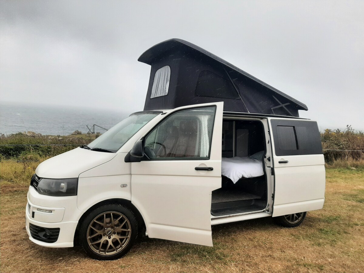 A white van is captured parked on grassy terrain, displaying its elevated pop-top roof. The sliding door is open, revealing a spacious interior with a bed setup. A scenic coastal backdrop can be seen in the distance, with cloudy skies above.