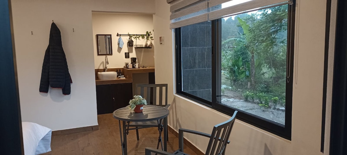 A cozy indoor seating area features a round table accompanied by two chairs. Natural light fills the space through a large window, revealing views of greenery outside. A small plant sits on the table, and items are neatly organized on a shelf nearby.