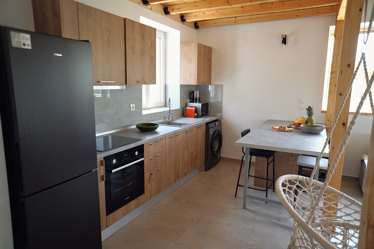A modern kitchen features wooden cabinetry and a sleek countertop. A black refrigerator and built-in oven are positioned alongside a washing machine. A dining table with two chairs is set with fruit, while light enters through a window, highlighting the functional kitchen space.