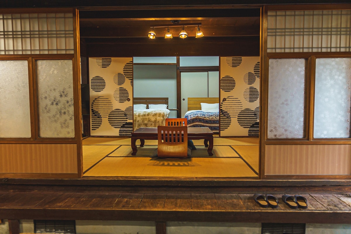 A traditional Japanese room is featured, showcasing two separate sleeping areas with comfortable beds. Sliding doors provide a view of tatami flooring, and a simple wooden chair sits in front of a low table. Natural light filters through the frosted windows.