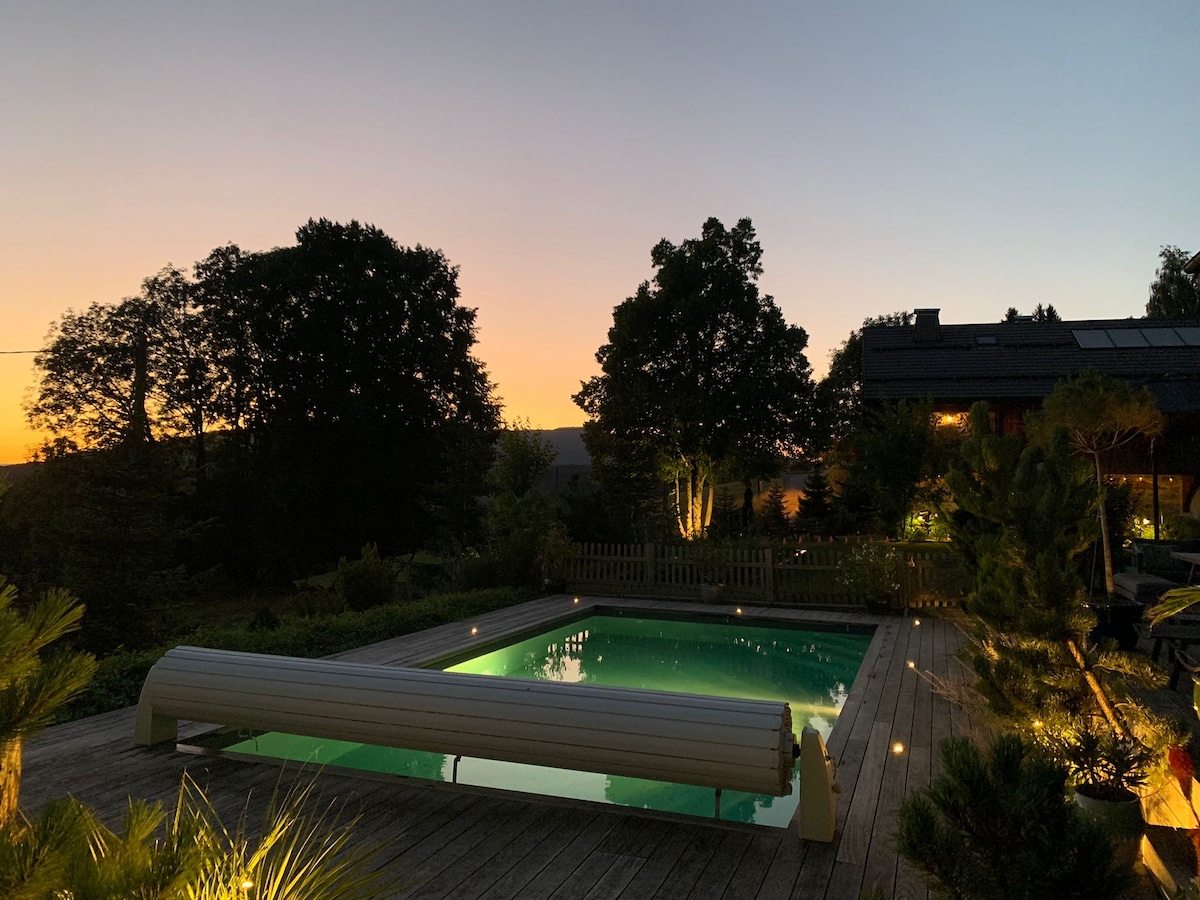 A serene outdoor pool is surrounded by a wooden deck, with ambient lighting creating a tranquil atmosphere. Lush trees frame the scene, while a sunset sky casts warm hues over the setting. The inviting pool is unoccupied, enhancing the feeling of peaceful solitude.