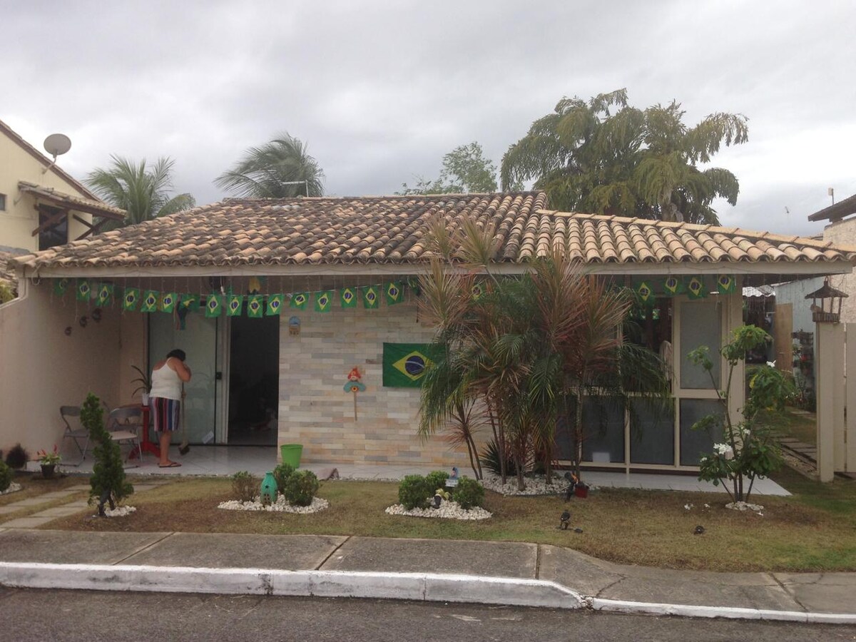 A single-story house features a tiled roof and decorative elements, including a Brazilian flag. Lush greenery surrounds the entrance, complemented by small plants and gravel accents in the yard. A person is seen engaging with the area, adding a sense of activity to the setting.