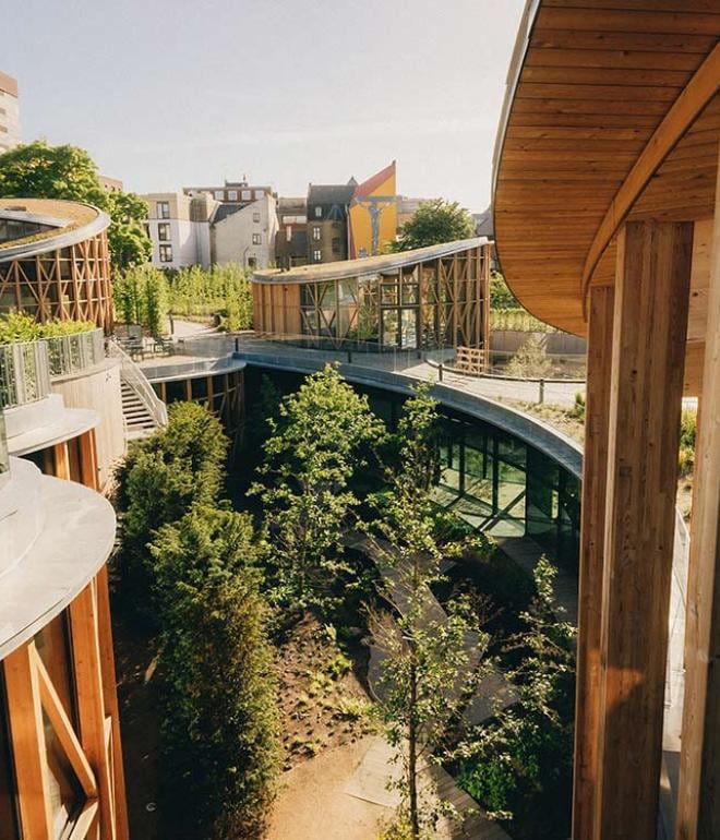 Curved wooden structures are interconnected, surrounded by lush greenery. Glass walls provide views of the landscaped areas, featuring small trees and pathways. Natural light fills the outdoor spaces, creating a harmonious blend of architecture and nature.