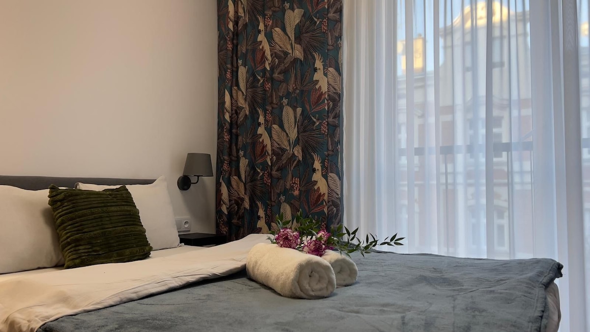 A well-appointed sleeping area showcases a spacious bed adorned with soft pillows and a textured green throw. Fresh flowers and rolled towels are placed on the bed, while large windows with sheer curtains allow natural light to fill the room.