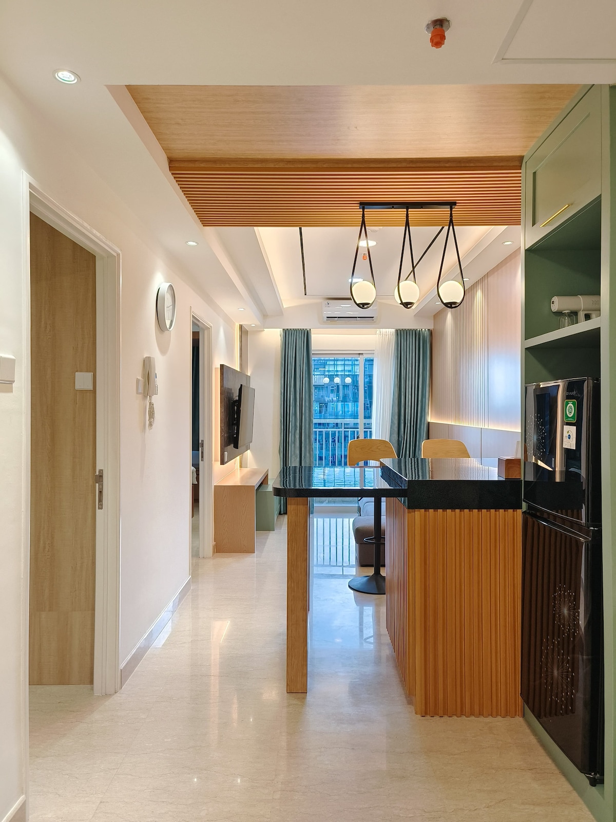 A modern kitchen area is highlighted by a wooden bar top with two wooden stools. Soft lighting is provided by a pendant lamp above. Natural light filters in through large windows, illuminating the open space, which includes a refrigerator and a wall-mounted television.