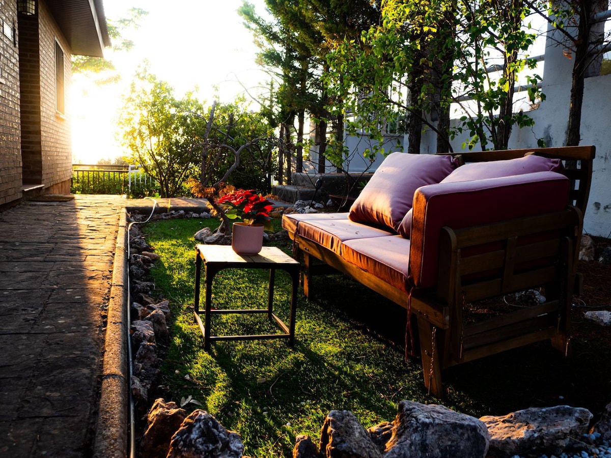 A cozy outdoor seating area is presented, featuring a wooden bench with plush cushions and a small table. Natural light filters through the surrounding trees, casting a warm glow on the grassy space, complemented by natural stone pathways.