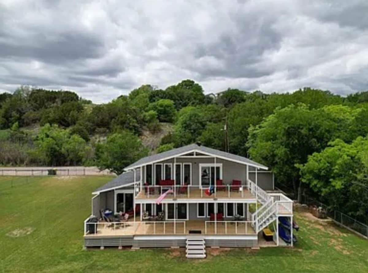 The lakeshore house features a spacious multi-level deck with seating and dining areas. Large glass doors provide access to the interior, while lush greenery surrounds the property. An expansive grass lawn leads to the house, creating a welcoming outdoor space.