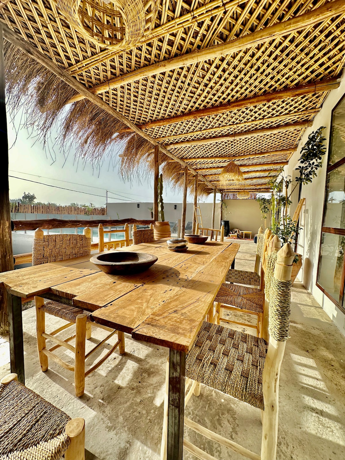 An outdoor dining area features a large wooden table surrounded by woven chairs. The area is shaded by a thatched roof, with sunlight filtering through, creating a relaxed atmosphere. Decorative bowls are placed on the table, enhancing the natural aesthetic of the space.