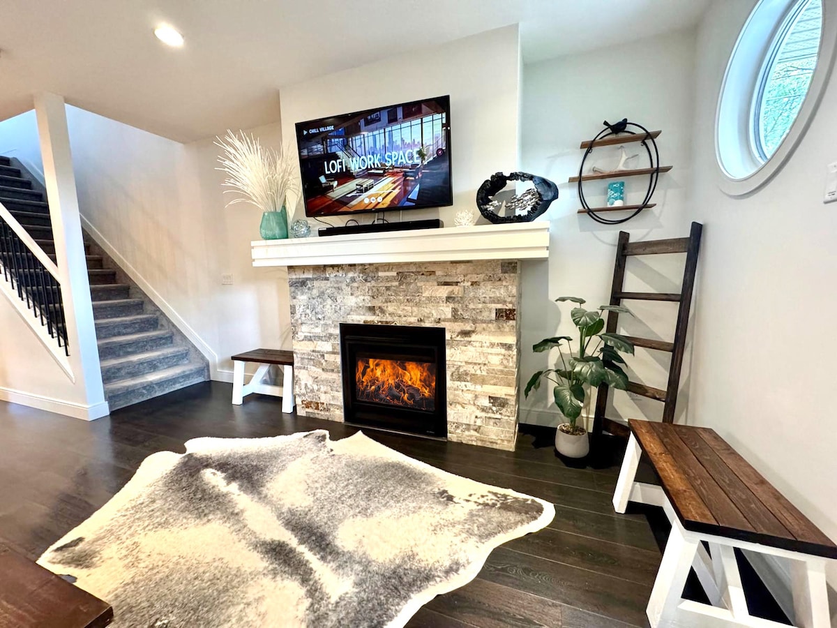 A modern living area features a stylish stone fireplace with a mounted television above it. Two wooden benches provide seating, with a decorative ladder nearby. A potted plant adds a touch of greenery, and a circular window allows natural light to enter the space.
