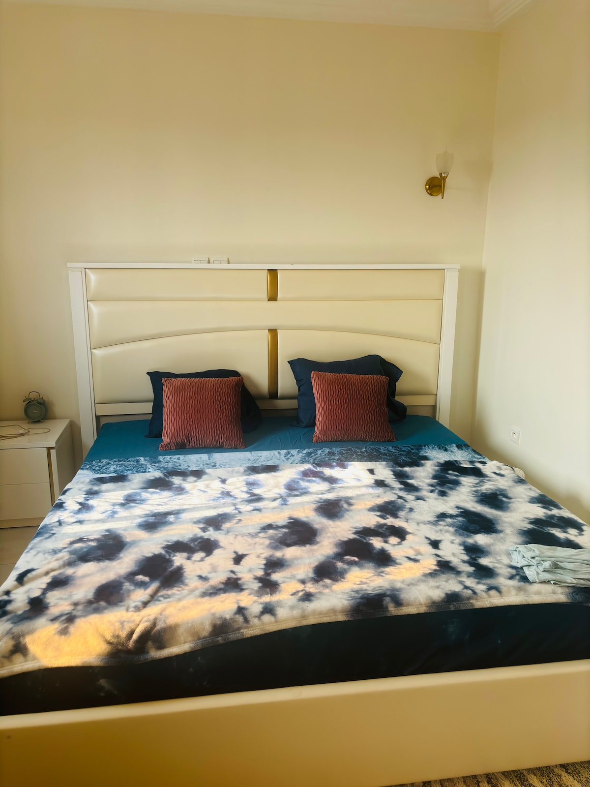 A spacious bed is dressed in a dark blue coverlet, accented by two contrasting decorative pillows in warm tones. A light-colored bedside table is positioned nearby, featuring a small lamp. Natural light gently fills the room, enhancing its calm ambiance.