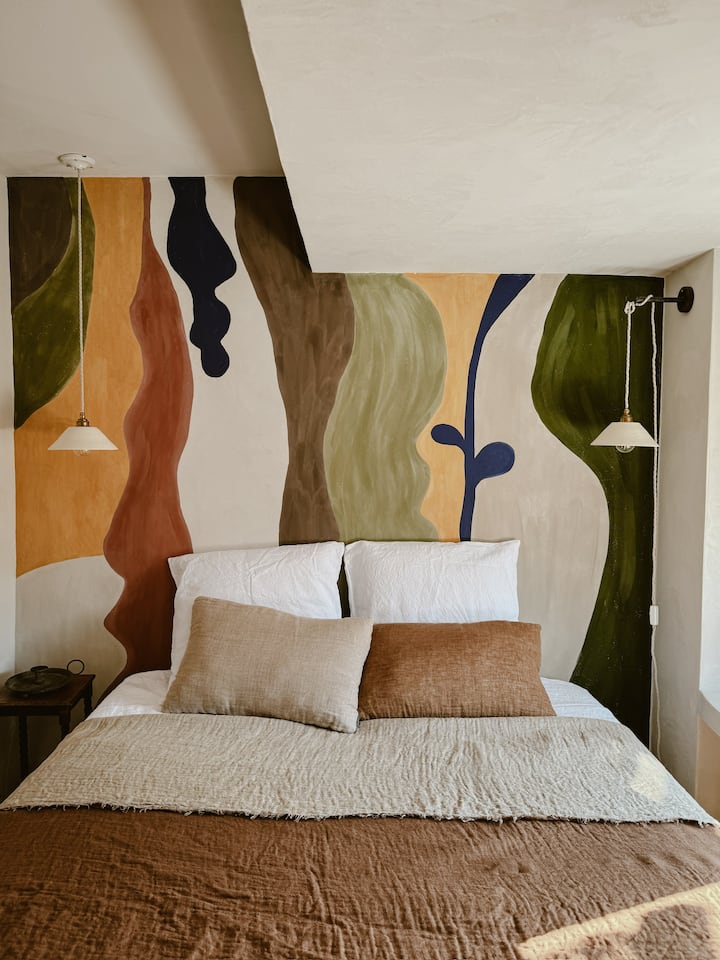 les murs recouverts d’un enduit rustique vous enveloppent et participent à l’atmosphère chaleureuse de l’espace. 

Sur la tête de lit, s’étend une fresque à la chaux naturelle, peinte à la main, évoquant les traits de l’artiste Rafael Uriegas.