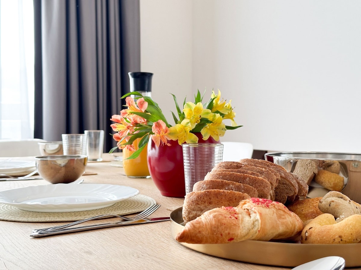 A dining table is set with plates and utensils, featuring an assortment of freshly baked pastries on a round tray. A vibrant bouquet of yellow flowers is displayed in a glass vase, accompanied by a pitcher of orange juice and additional glassware.