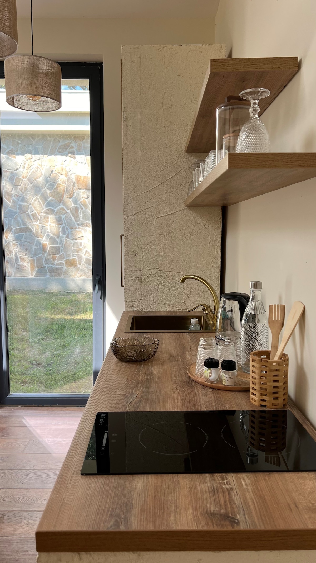 A well-appointed kitchen area features a smooth wooden countertop with an integrated stovetop. Open shelving displays glassware and utensils against a light wall. Large windows allow natural light to illuminate the space, while greenery is visible just outside.