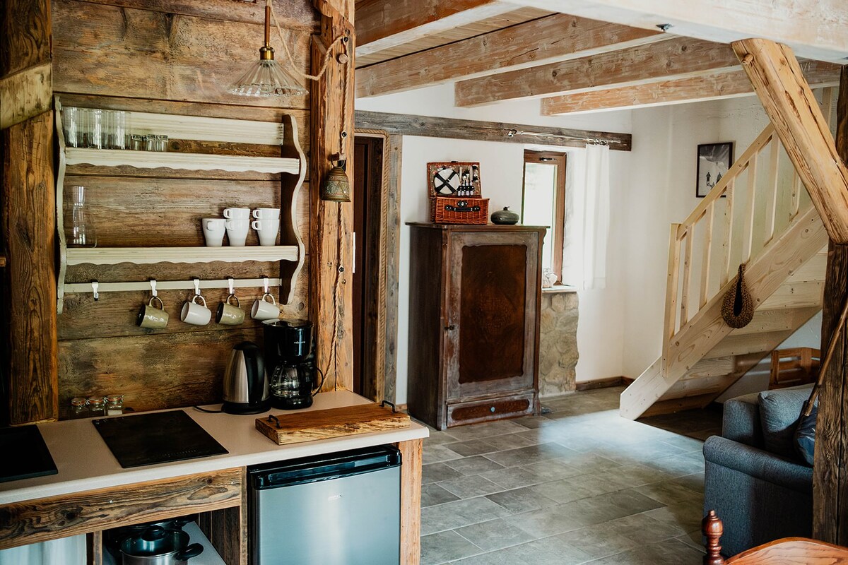 An open living area features rustic wooden beams and a stone wall, enhancing the cozy ambiance. A compact kitchen with a shelf displays mugs and glassware, while a staircase leads to the upper floor. Natural light fills the space from nearby windows.