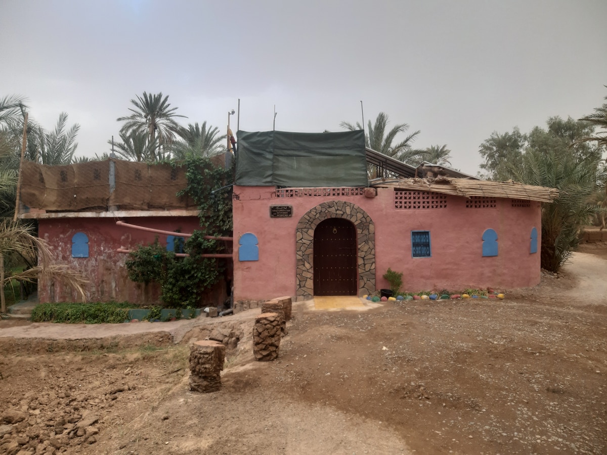 A charming pink building is seen, featuring a stone arched entrance and blue window shutters. A thatched roof and lush greenery enhance the natural setting. The surrounding area includes palm trees and an unpaved path, creating a tranquil outdoor environment.