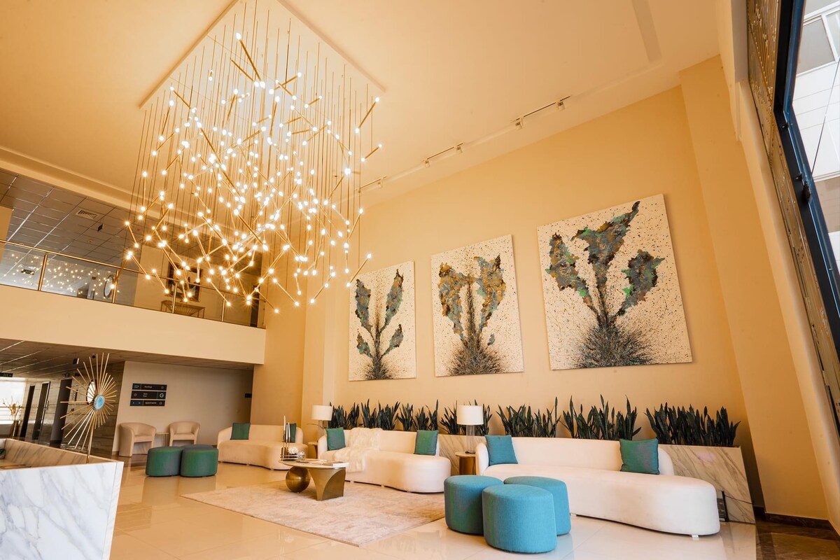 The lobby space is characterized by high ceilings and modern lighting fixtures. Textured wall art depicting abstract plant forms is displayed prominently. A comfortable seating area is arranged with circular ottomans and plush couches, providing a welcoming environment.