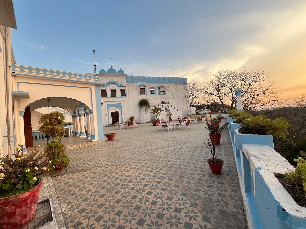 A wide terrace area is visible, adorned with intricate tile patterns and potted plants. The background features a heritage structure with blue accents and multiple arched doorways. A sunset casts a warm glow over the scene, enhancing the tranquil atmosphere.