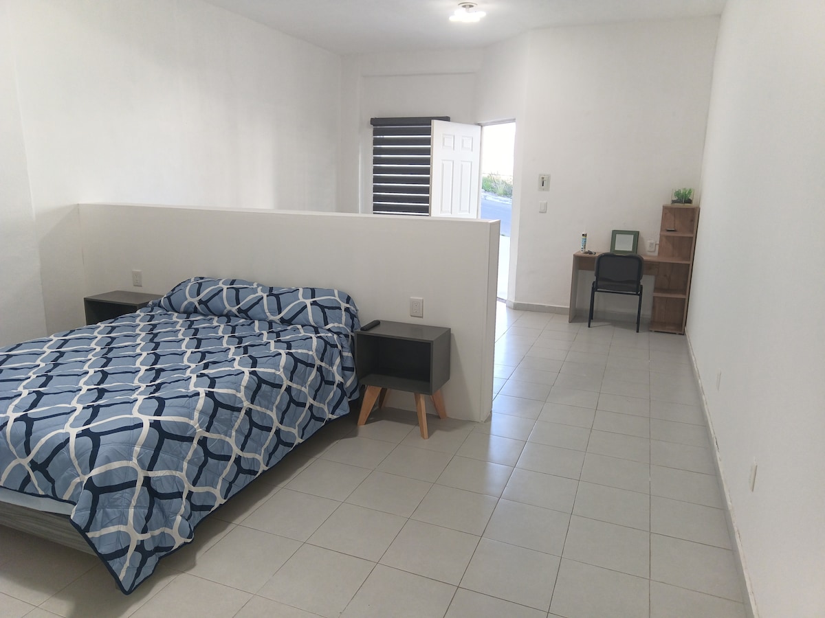 A spacious loft area is depicted, featuring a neatly arranged Queen size bed with a geometric patterned comforter. Nightstands are positioned on either side of the bed. A work desk and chair are visible, along with a window allowing natural light into the room.