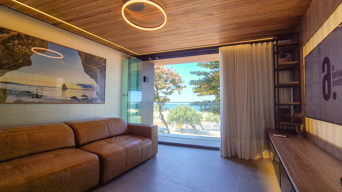 A living area is showcased with a large window offering a view of greenery and the beach. A leather sofa sits opposite a wooden desk, while art pieces adorn the walls. Soft lighting is provided by a circular fixture above, complemented by wooden ceiling panels.