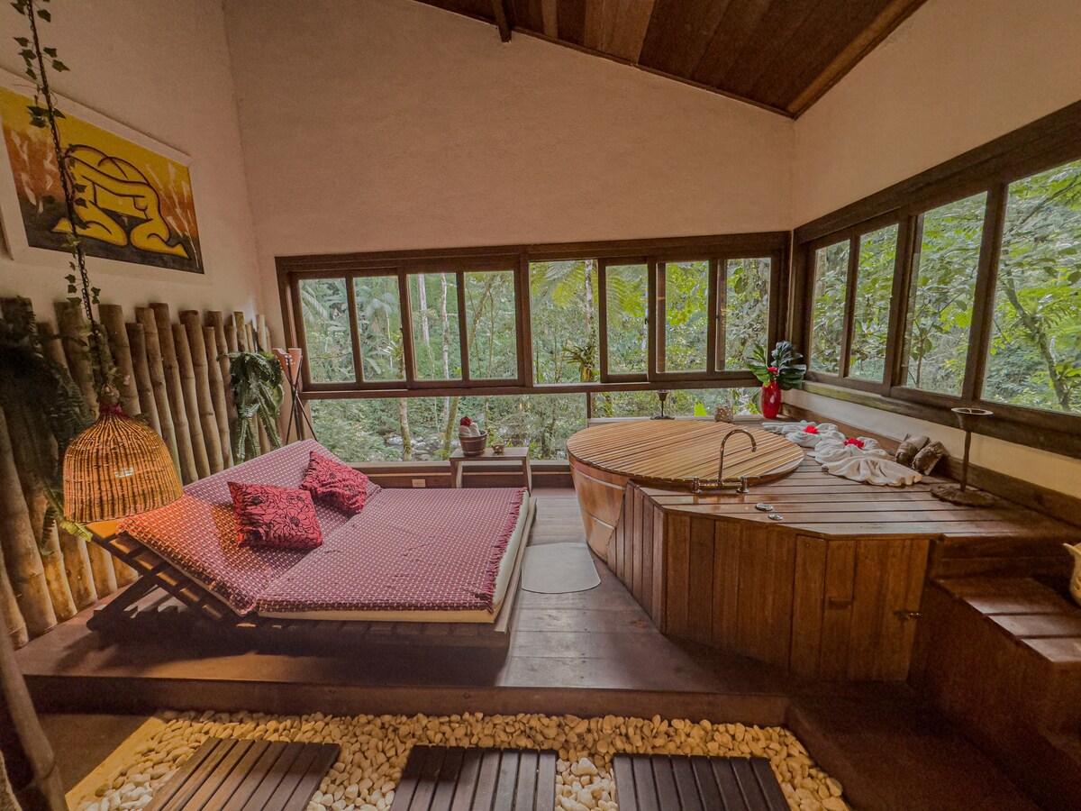 A cozy interior features a spacious bed with patterned bedding positioned alongside a wooden hot tub. Large windows provide views of the surrounding greenery, and natural wood accents enhance the connection with nature, creating a serene atmosphere.