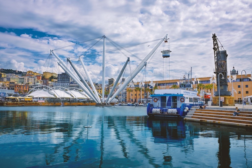 The image captures the vibrant Porto Antico area of Genoa, featuring a striking modern structure with large white cables arching overhead. Reflections on the calm water highlight the blue sky, while boats are visible in the foreground, enhancing the scene's lively atmosphere.