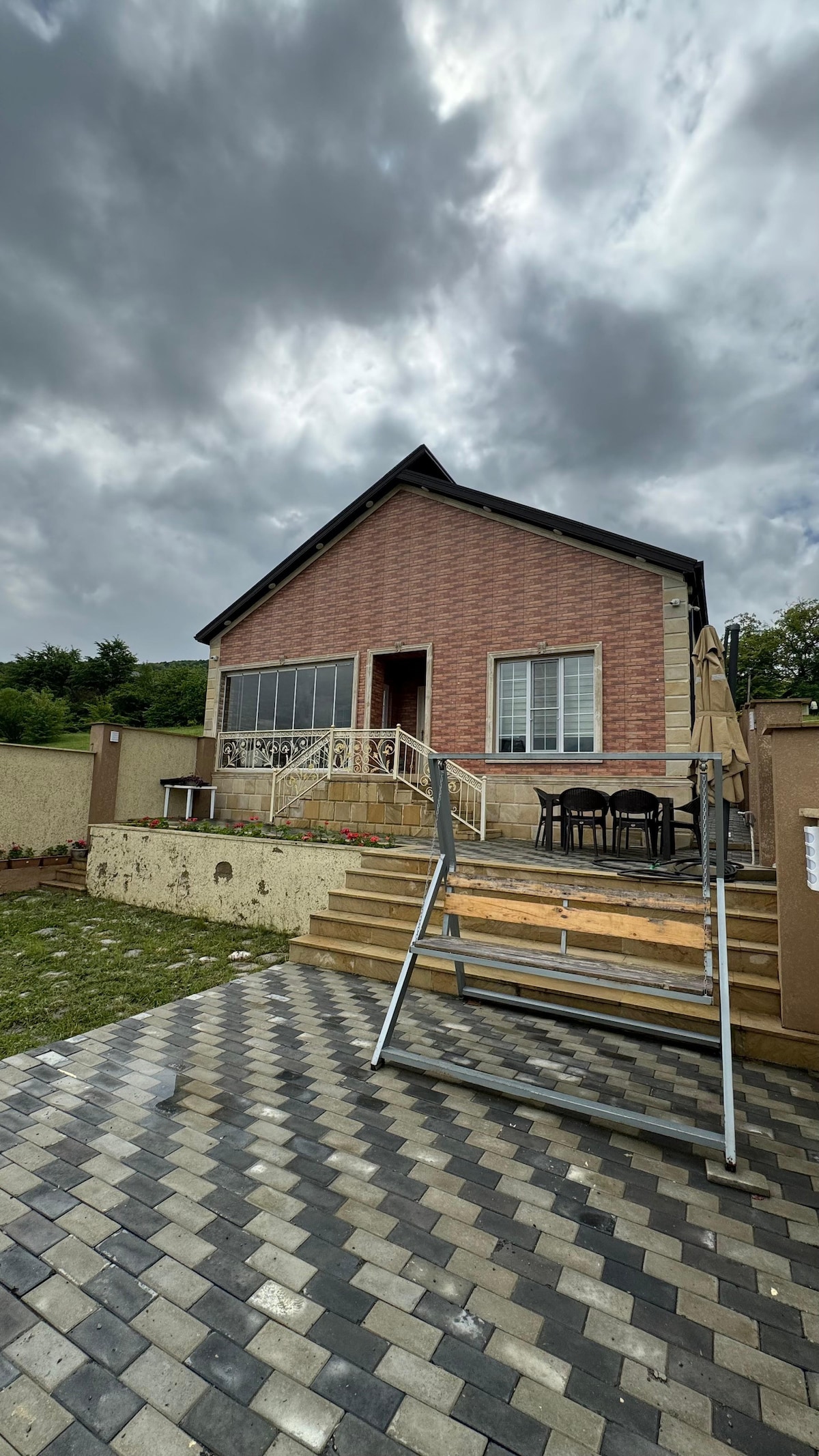 A charming house exterior is captured, showcasing a brick façade under a cloudy sky. A wooden deck is accessed by a short set of stairs, with outdoor seating arranged nearby. The surrounding area features a patterned stone pathway leading to a grassy lawn.