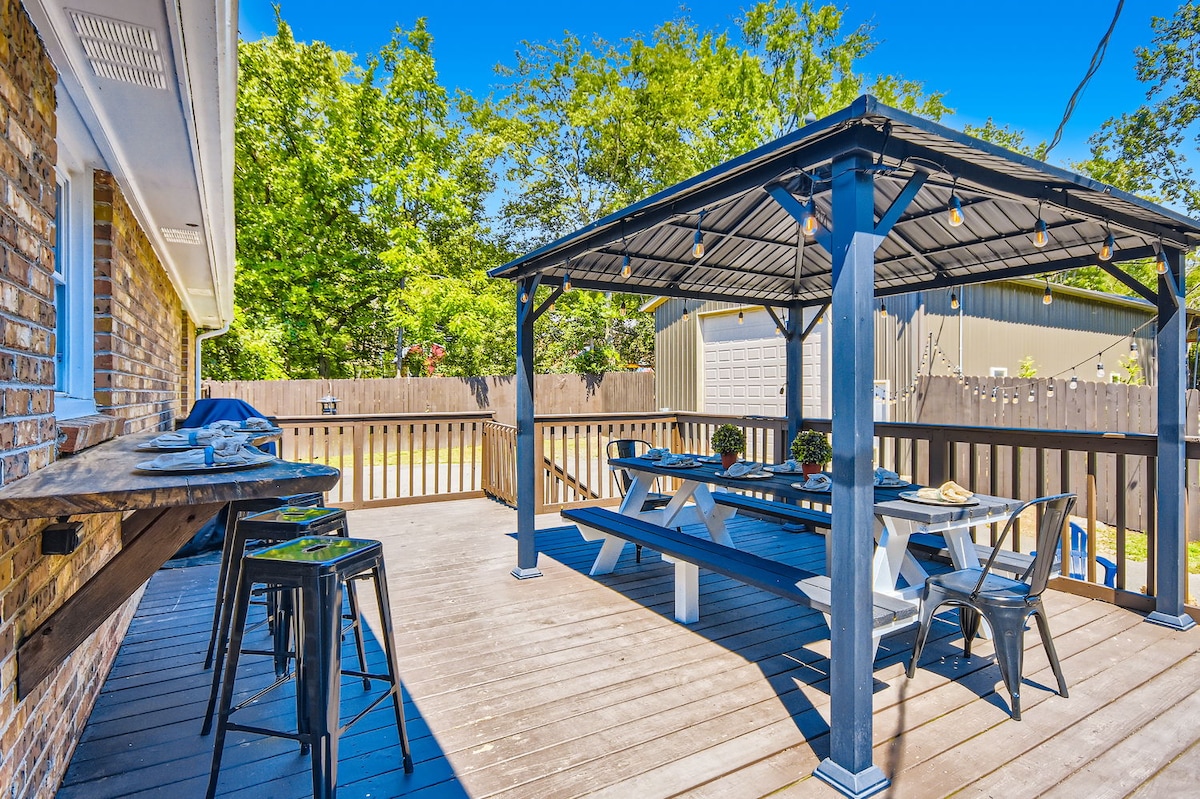 An outdoor patio area is presented, featuring a large wooden table with ample seating beneath a sheltering gazebo. Additional bar seating is positioned against a side railing, while string lights enhance the space. Lush greenery surrounds the patio, contributing to a tranquil ambiance.