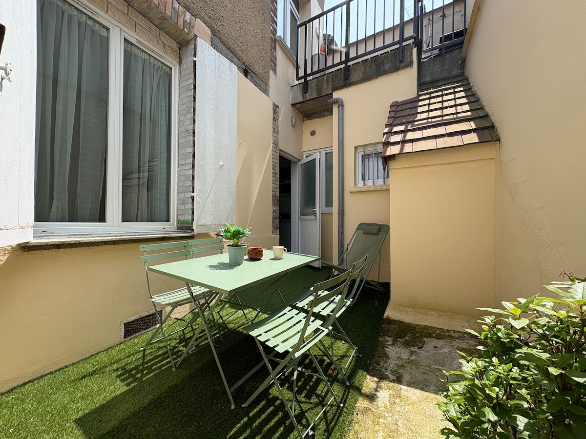 A private terrace area is furnished with a green table and several chairs. Sunlight filters through nearby windows, creating a bright space. Lush greenery borders the space, providing a serene outdoor setting for relaxation. A small building structure is visible in the background.