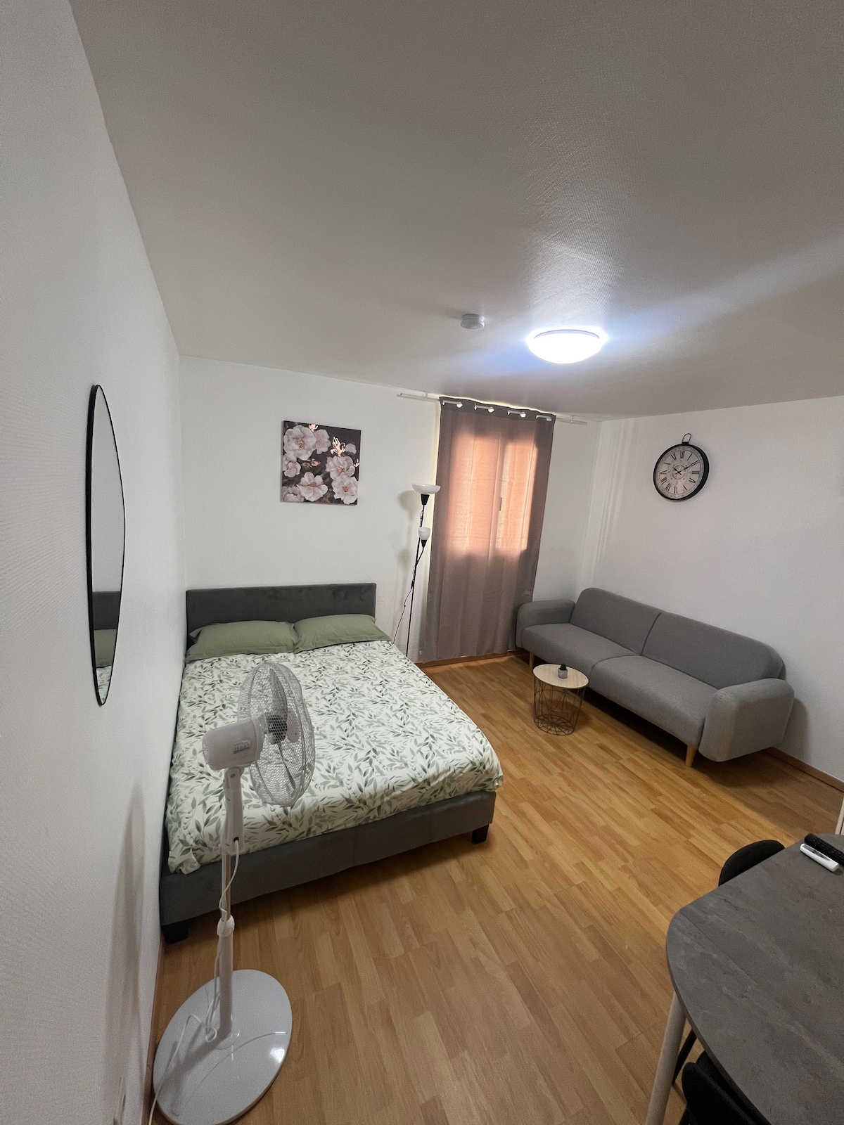 The room is furnished with a spacious bed topped with a floral-patterned blanket. A gray sofa is positioned across from a small round table. Light wood flooring complements the neutral walls. A mirror and wall clock add functionality, while curtains frame the window, filtering natural light.