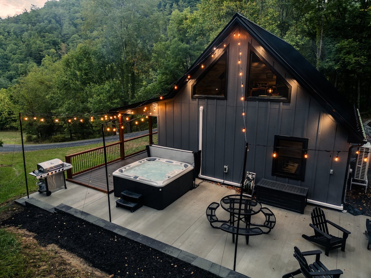 An outdoor area is viewed from above, featuring a hot tub surrounded by string lights that create a soft glow. A grill is positioned nearby on a patio surface, while the cabin's modern design is complemented by natural greenery in the background.