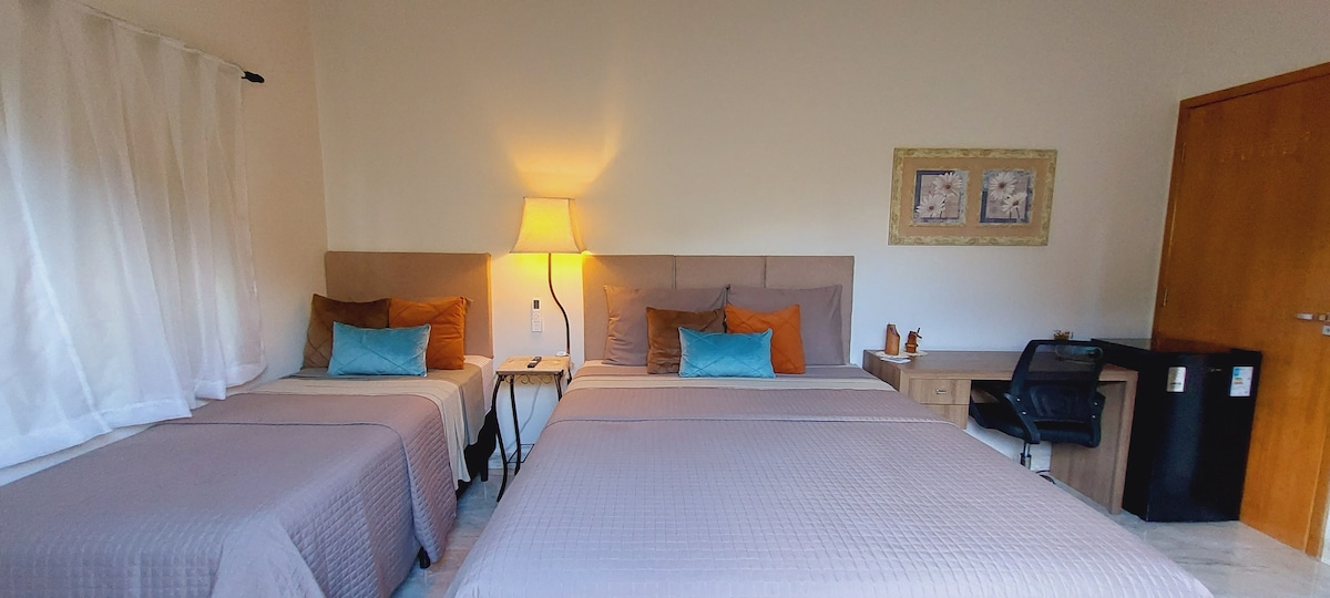 The sleeping area features a queen bed alongside a single bed, both dressed in light bedding. A bedside table with a lamp provides soft illumination. Adjacent, a workspace with a chair and a small refrigerator is visible, contributing to the room's functionality.