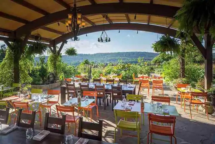 An outdoor dining area features a canopy overhead, with a variety of colorful chairs in orange and yellow surrounding wooden tables. Lush greenery and planters enhance the space, while a scenic view of rolling hills is visible in the background under a clear sky.