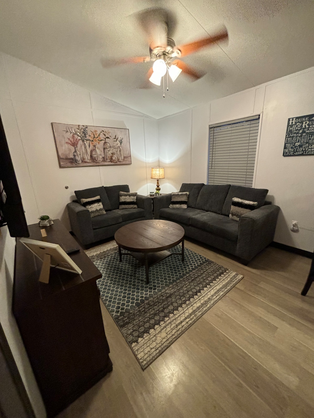 A comfortable living area features two gray sofas facing each other, adorned with decorative cushions. A round wooden coffee table sits at the center, surrounded by a patterned area rug. Soft lighting is provided by a standing lamp next to a window with blinds.