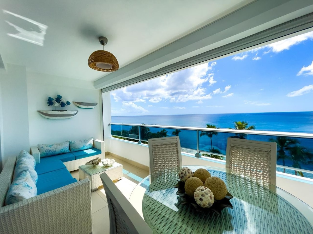 A bright living space features a comfortable seating area with a light blue sofa, complemented by a round glass dining table set. Large windows provide unobstructed views of the ocean and sky, enhancing the airy atmosphere with natural light.