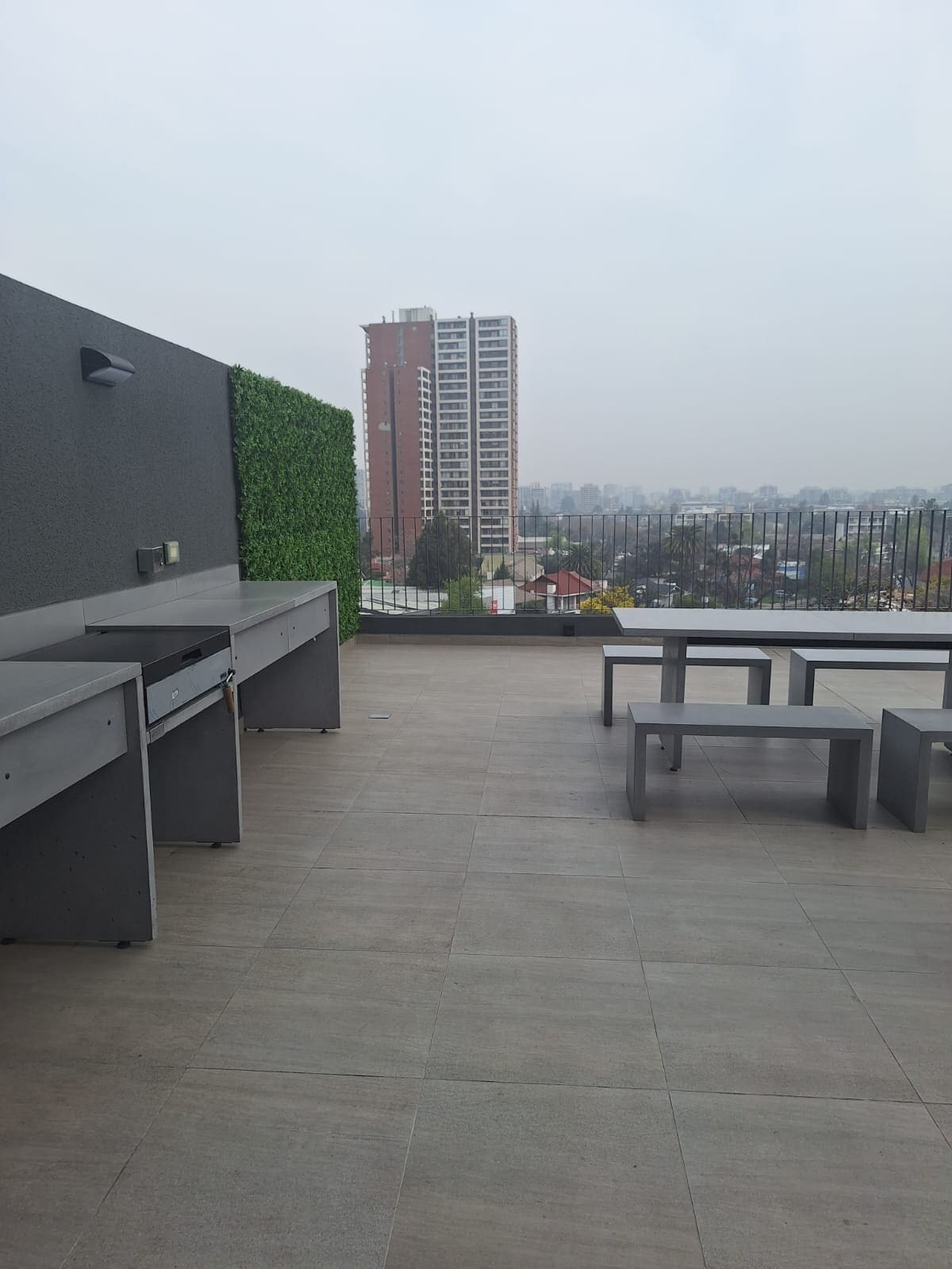 A rooftop space features modern gray outdoor furniture, including a table and chairs. A barbecue area is positioned against a wall with green foliage, providing a contrast to the urban backdrop. Distant city buildings and a hazy skyline create a serene atmosphere.