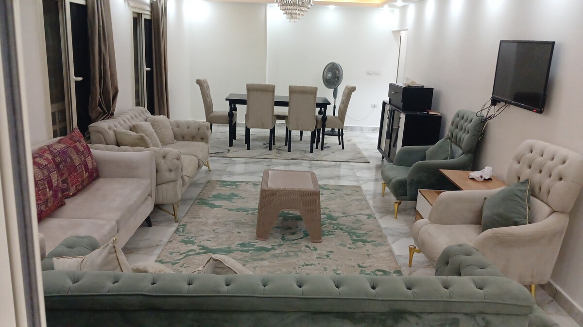 An elegant living area is presented with plush seating, including a pair of soft green sofas, upholstered chairs, and a light-colored rug. A dining table with six chairs is positioned nearby, while a flat-screen television and a fan are visible on the wall.